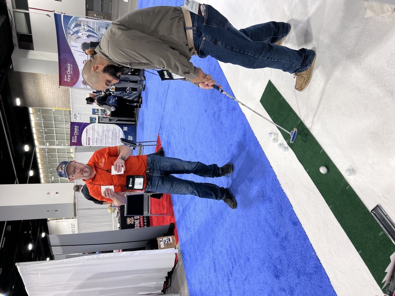 Man putts golf ball on mini-green at a trade show. Another man watches and gestures.
