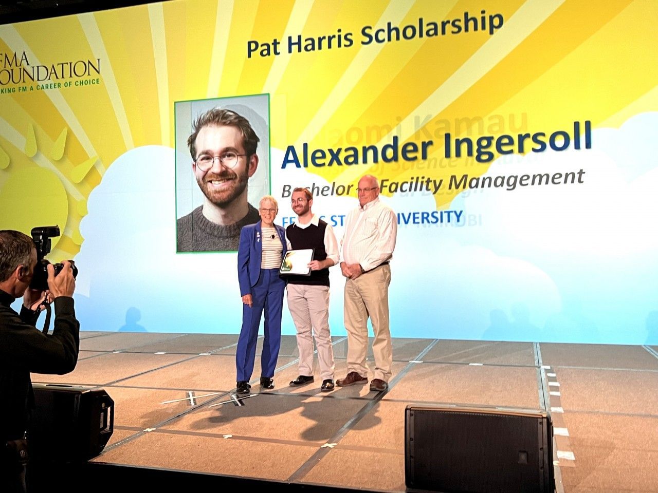 Man receiving scholarship on stage with two others. Backdrop: Pat Harris Scholarship, clouds, sun rays.