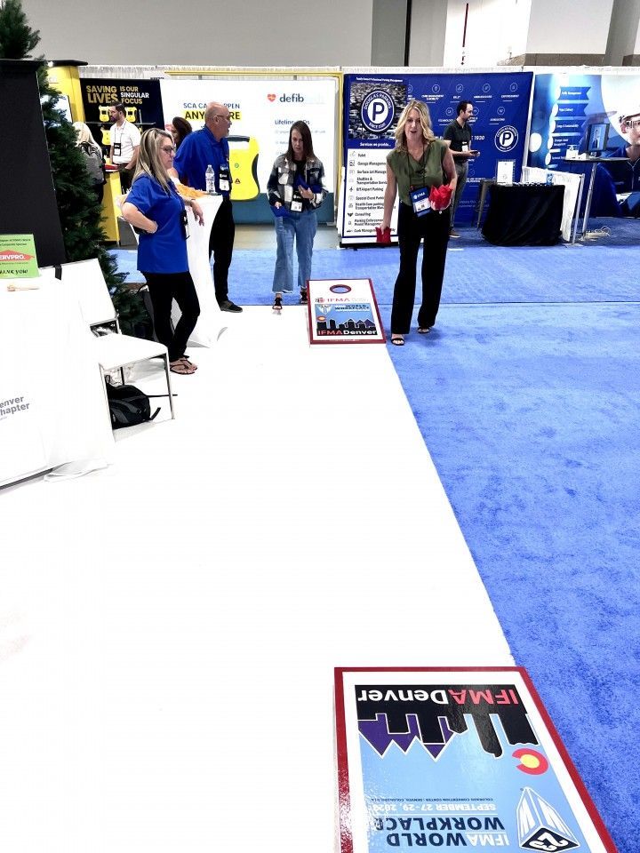 People playing cornhole at a trade show booth, on a white walkway with blue carpeting.