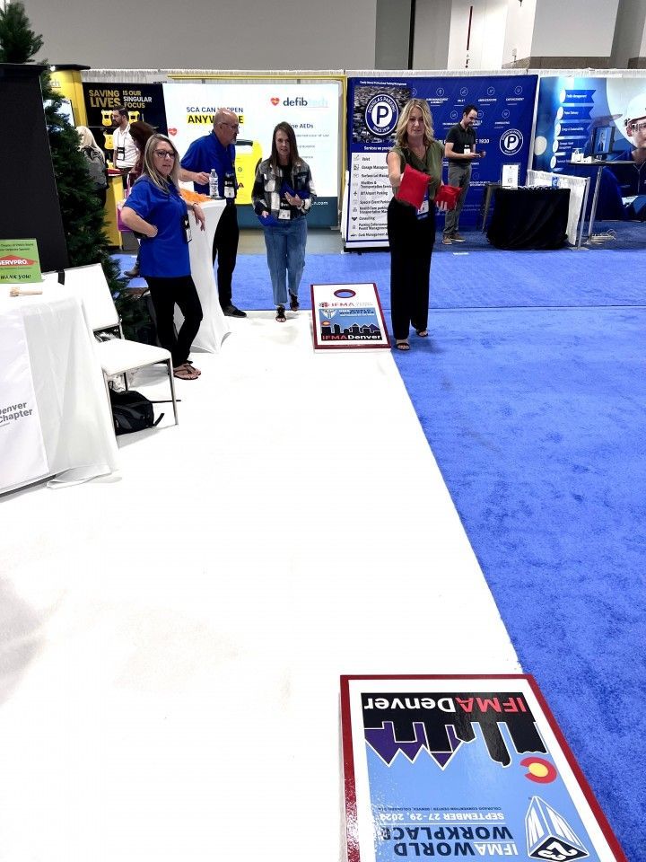 People playing bean bag toss at a booth, likely at a conference. Indoor setting with booths and carpet.