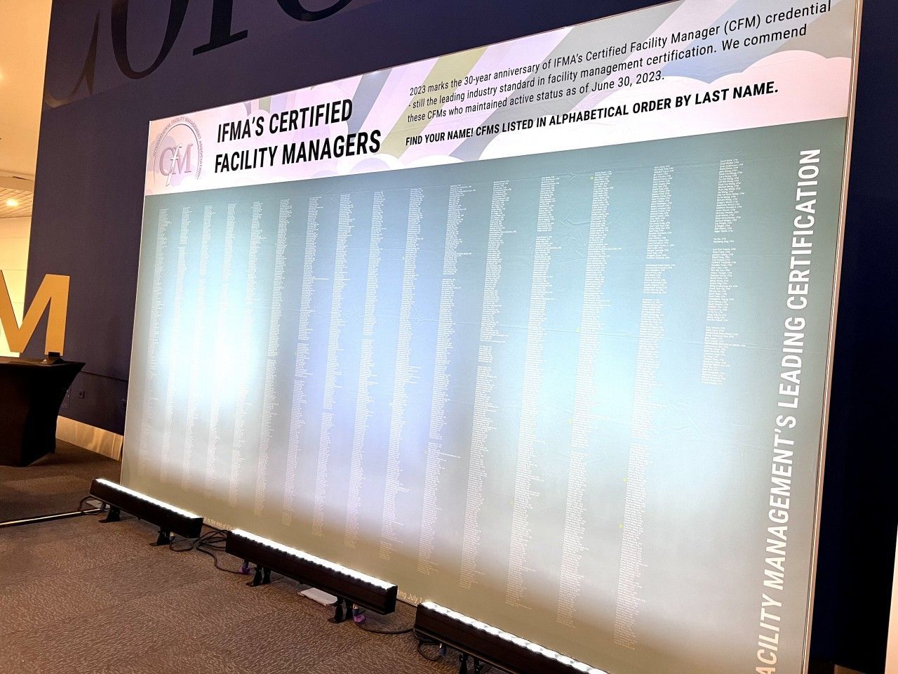 A large sign listing certified facility managers, mounted on a wall and illuminated by spotlights.