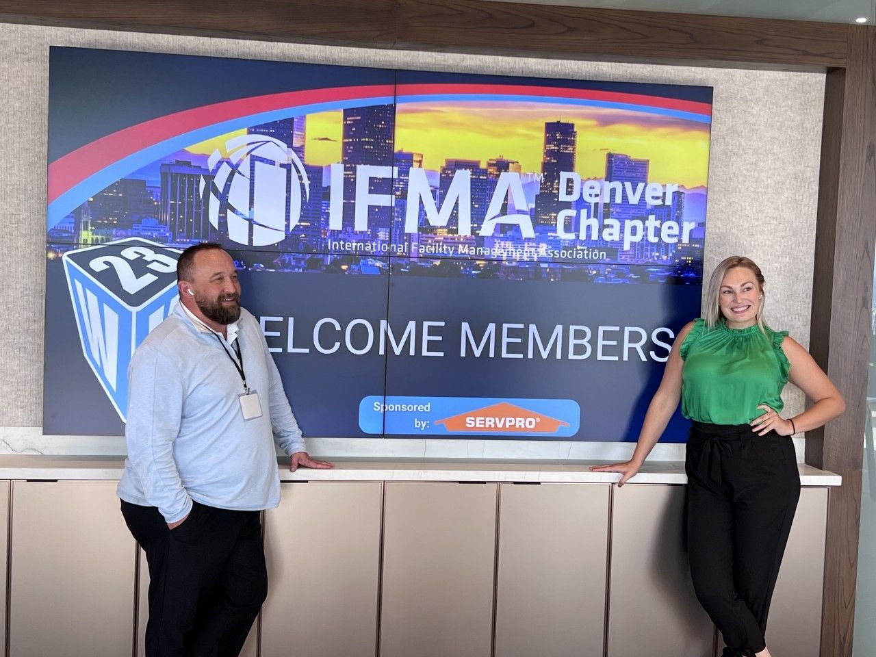 Two people stand by an IFMA Denver Chapter welcome sign. Man in blue shirt, woman in green top.