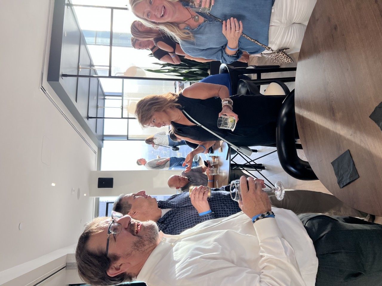 People socializing at a restaurant, seated around tables. Sunlight streams in through the windows.