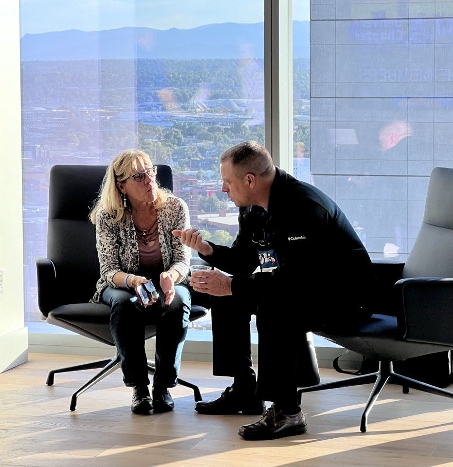 Two people sit in modern chairs, conversing in an office with a city view.