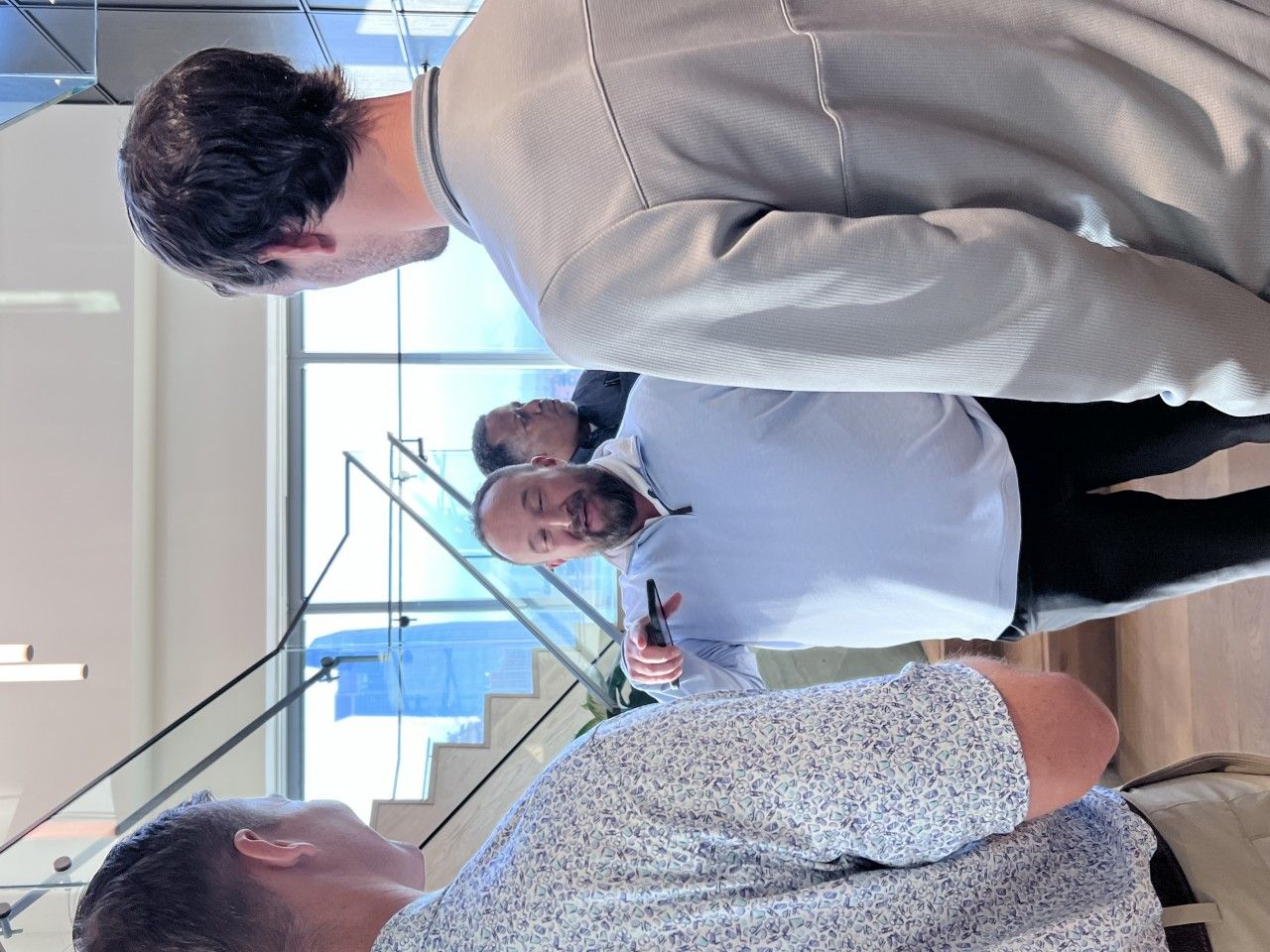 Men in business attire conversing near a staircase and windows. One man gestures with a pen.