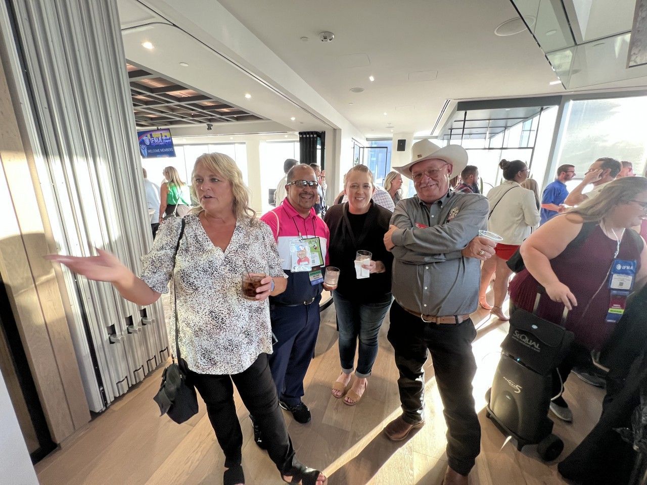 People in a social setting, woman gesturing, man in cowboy hat, other attendees. Interior, natural light.