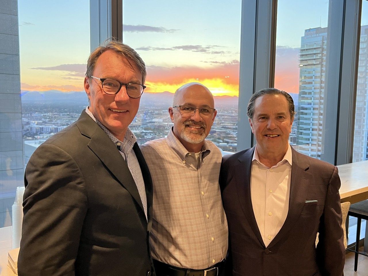 Three men in suits pose in front of a window overlooking a city skyline at sunset.