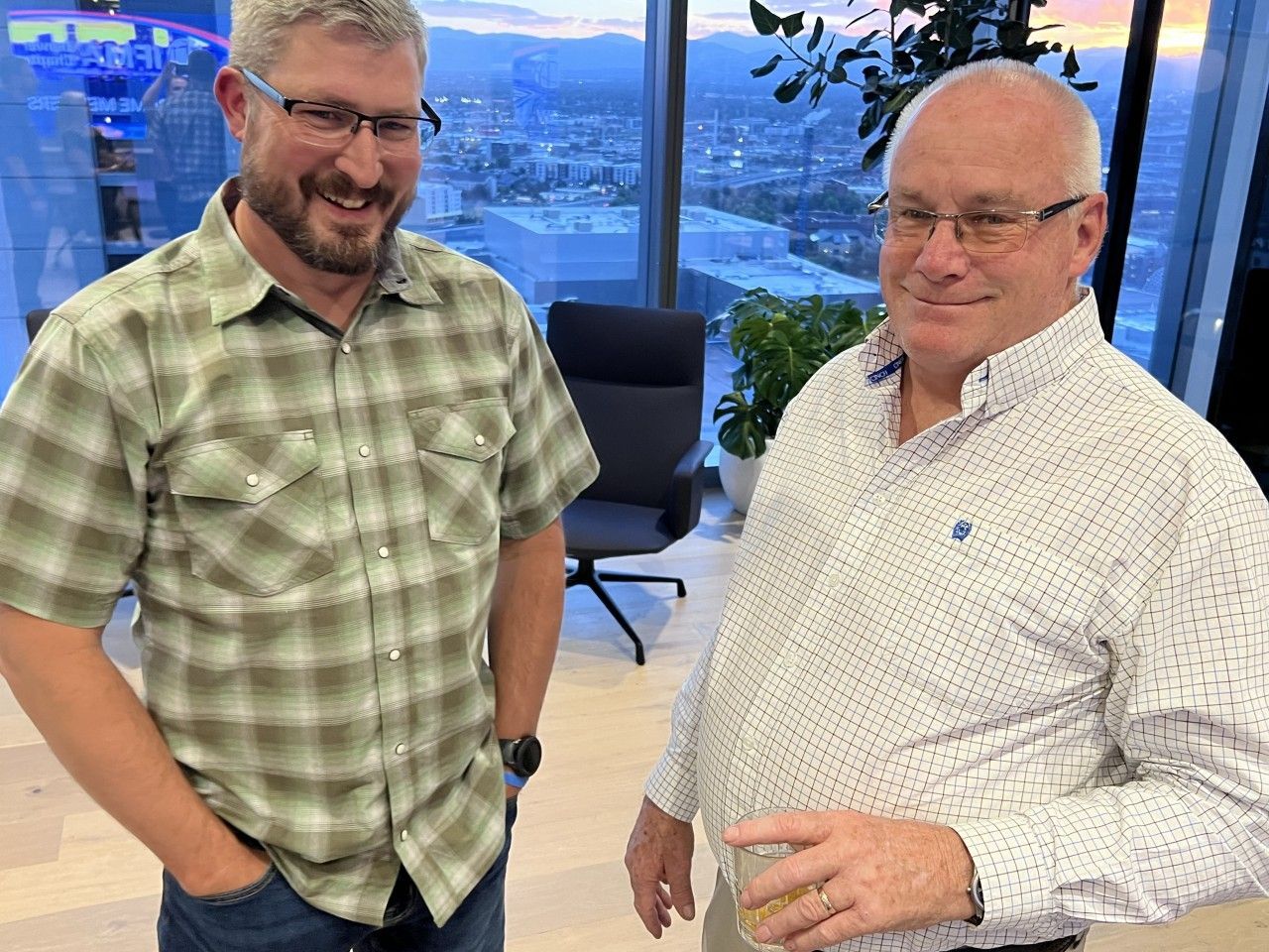 Two men smiling indoors, standing by a large window with city views.
