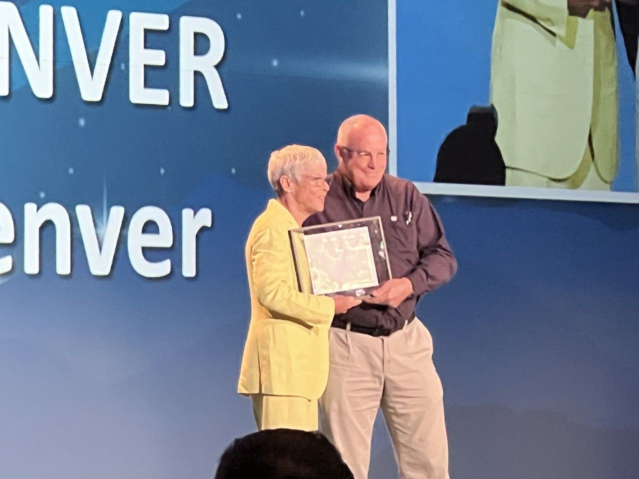 Woman in yellow suit presents framed document to man in grey shirt. Denver sign in background.