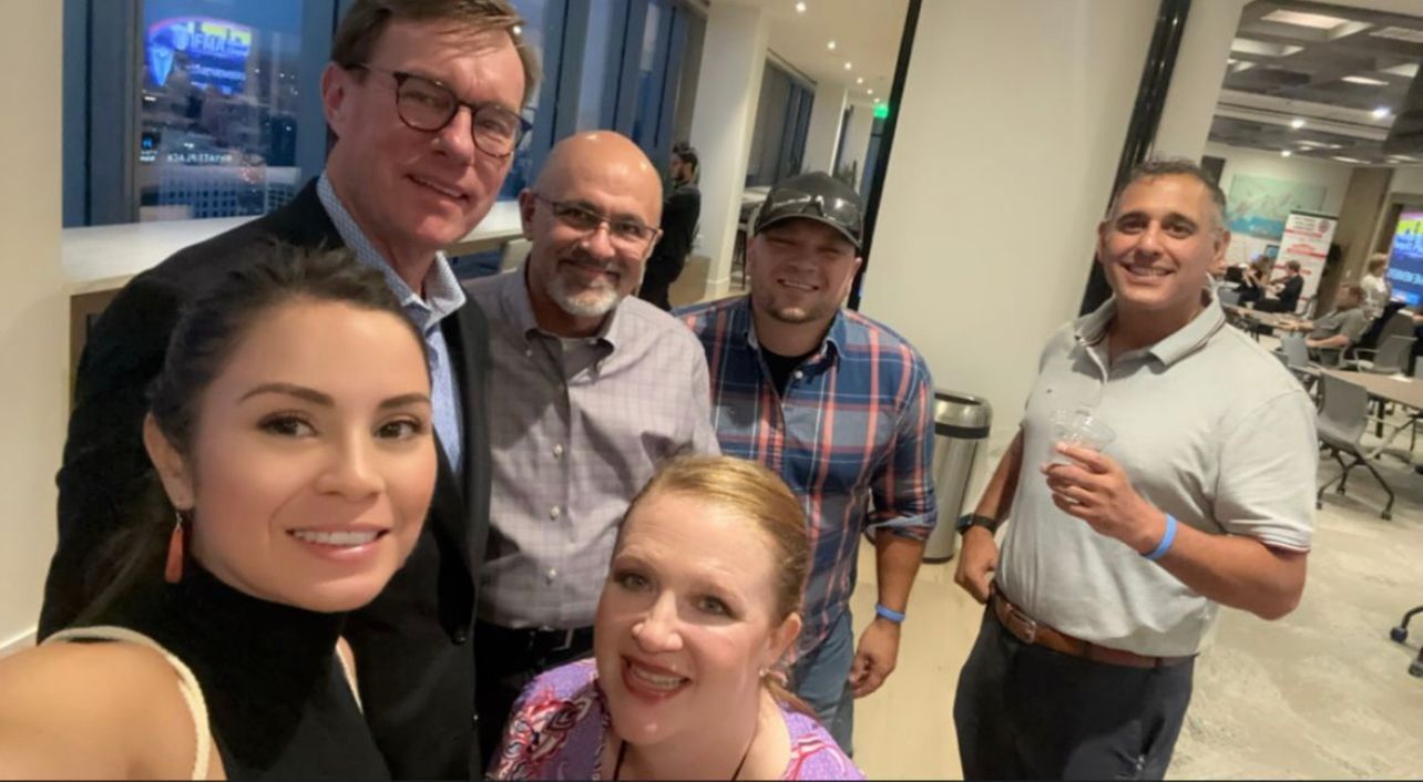 Group selfie: Six people smiling indoors. One woman holds the camera. Lighting is bright.