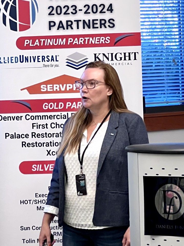 Woman speaking at a presentation, standing near a podium with partner logos in the background.