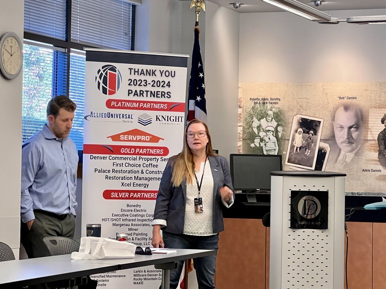 A woman speaks at a podium in an office, a man stands beside a banner listing sponsors.