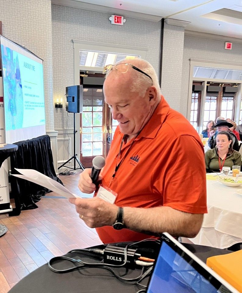 Man in orange shirt speaking at a conference, holding papers and a microphone. A screen and laptop are visible.