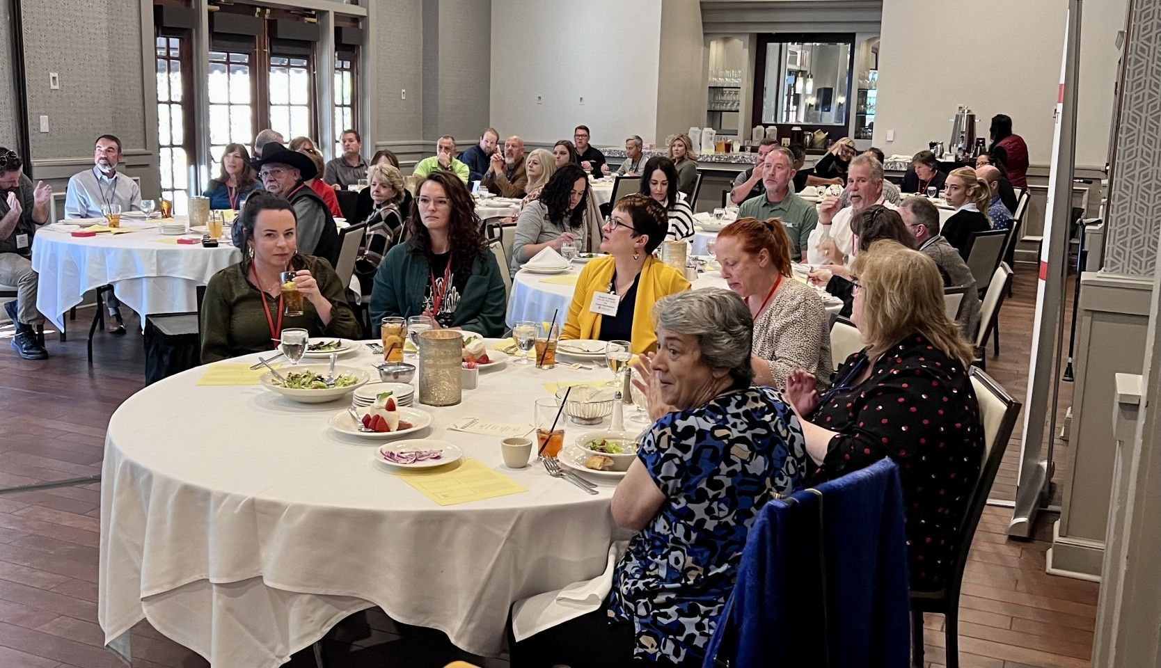 People seated at round tables in a banquet hall, some clapping. White tablecloths, food visible.