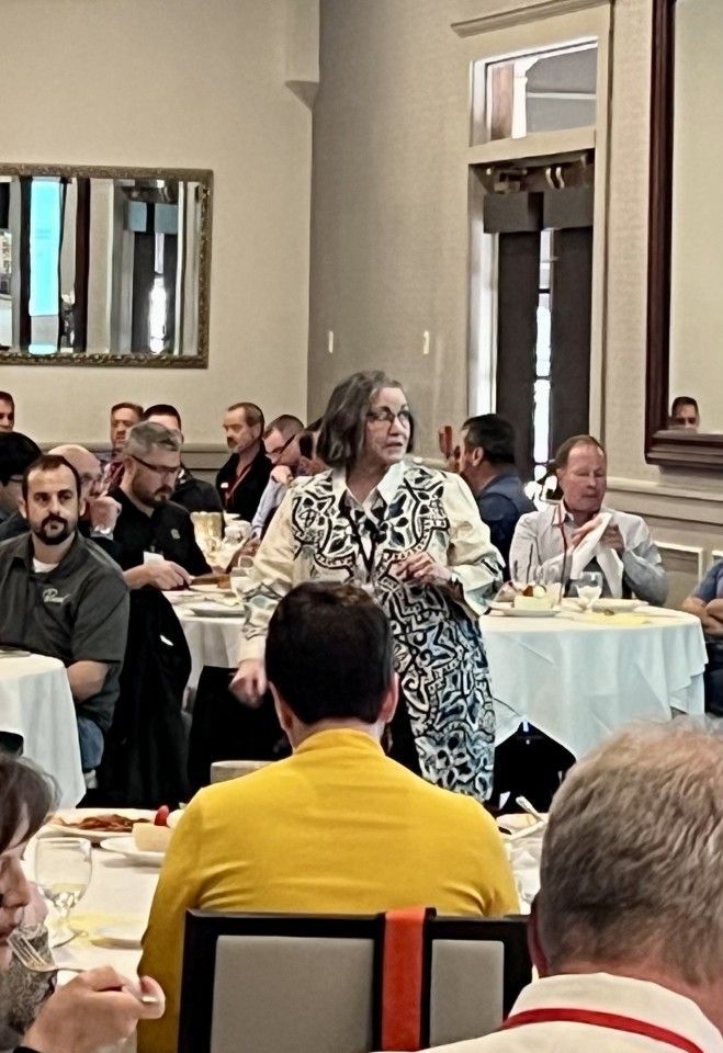 Woman speaking at a conference. People seated at round tables in a dining hall.