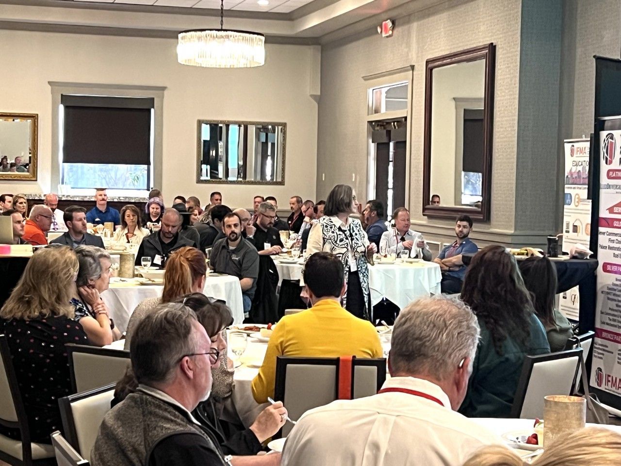 People seated at round tables in a conference room, listening to a presentation.