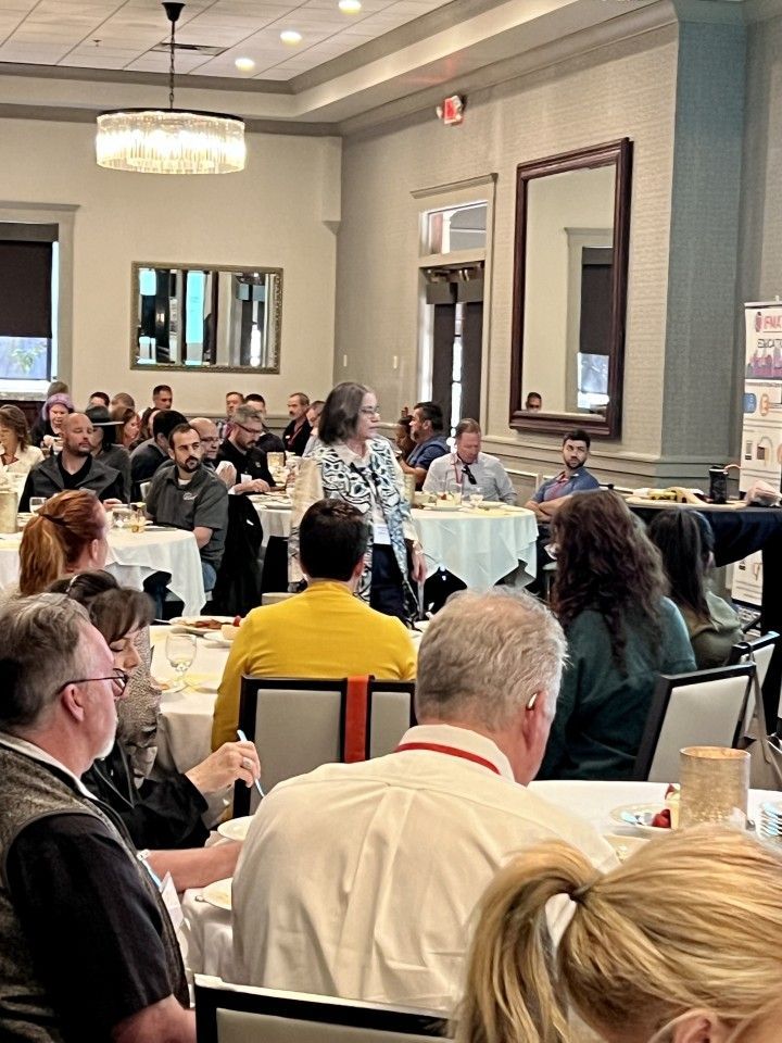 Woman speaking to a seated audience at tables in a brightly lit banquet hall.