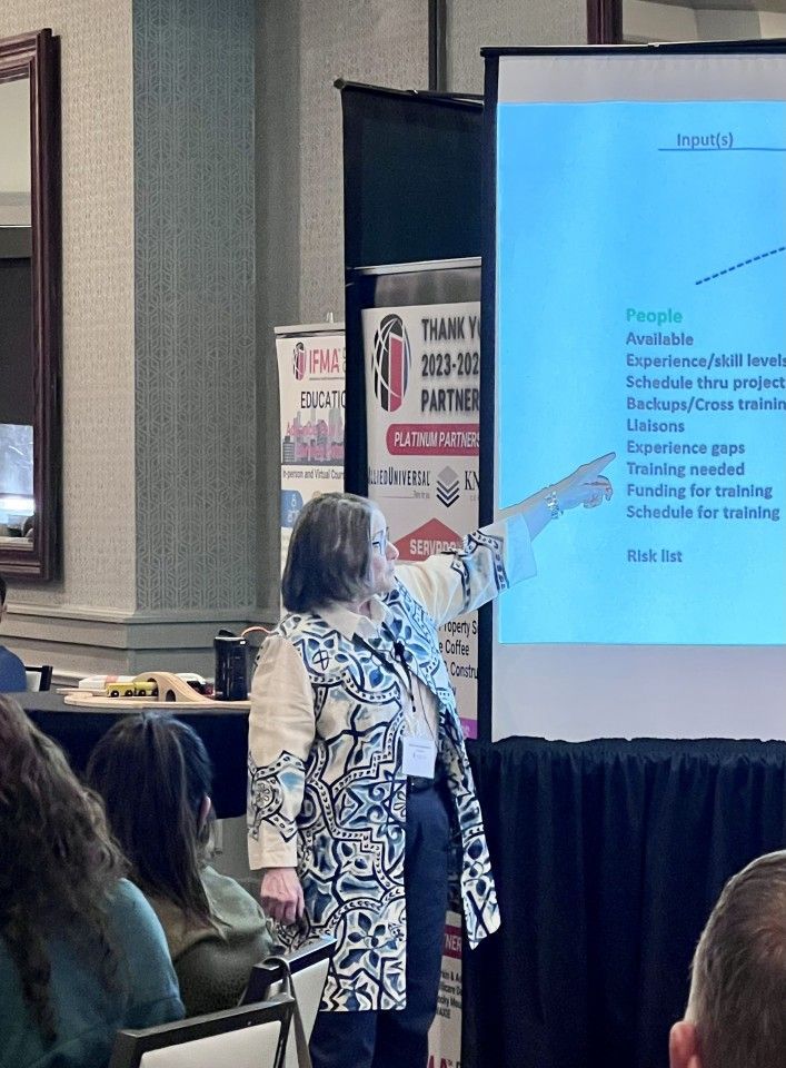Woman pointing at a presentation screen during a conference. Audience sits in foreground.