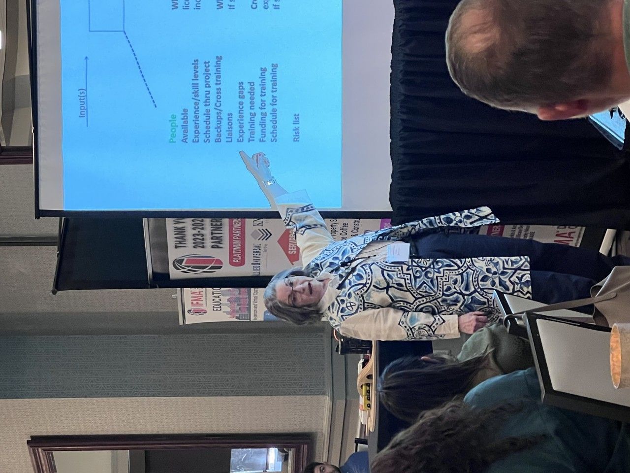 Woman presenting at a conference, pointing to a slide with text. People in the audience.