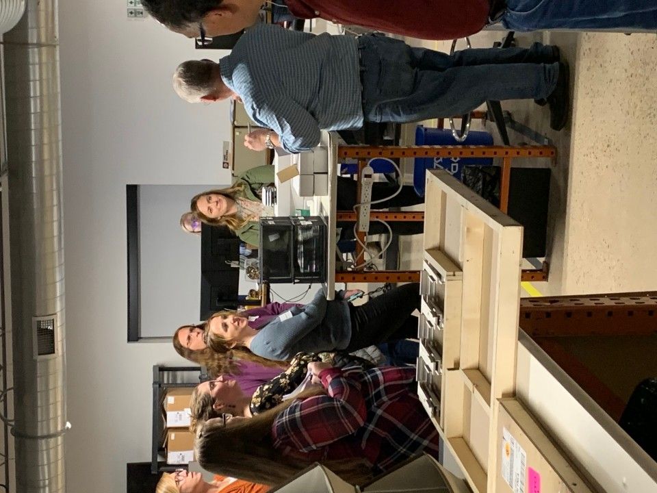 Group of people listening to a man speaking, possibly a presentation. Indoor setting with lab equipment.
