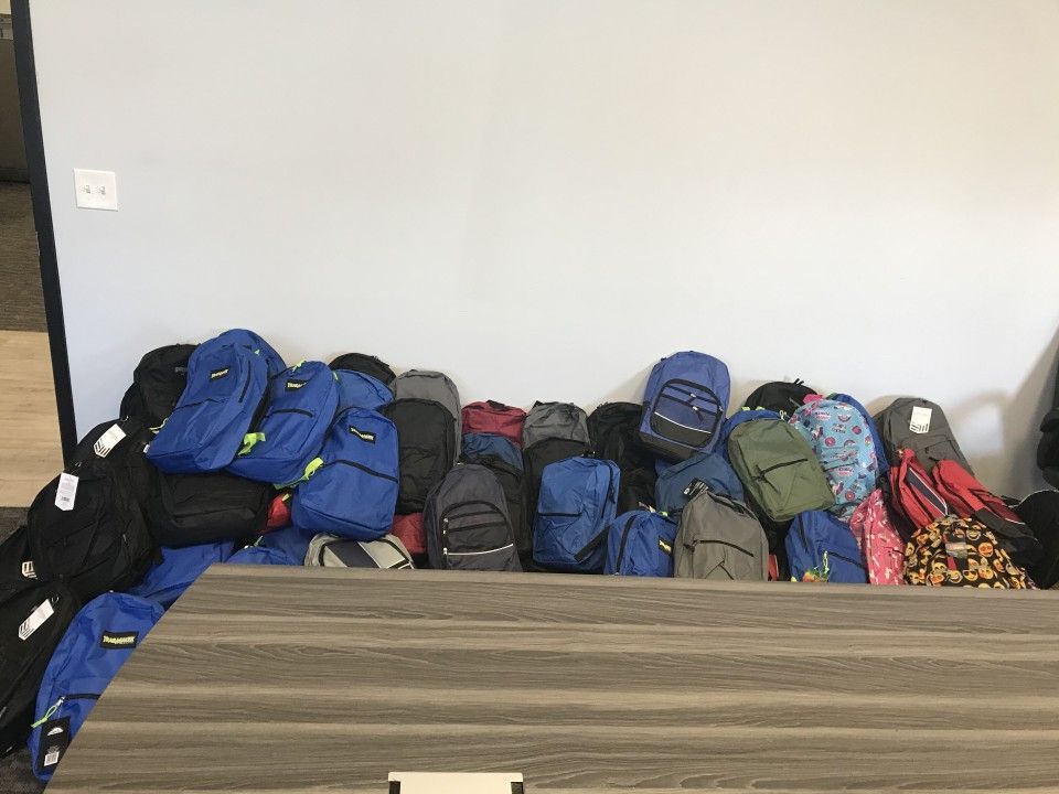 Pile of backpacks in various colors against a gray wall, on a wooden surface.