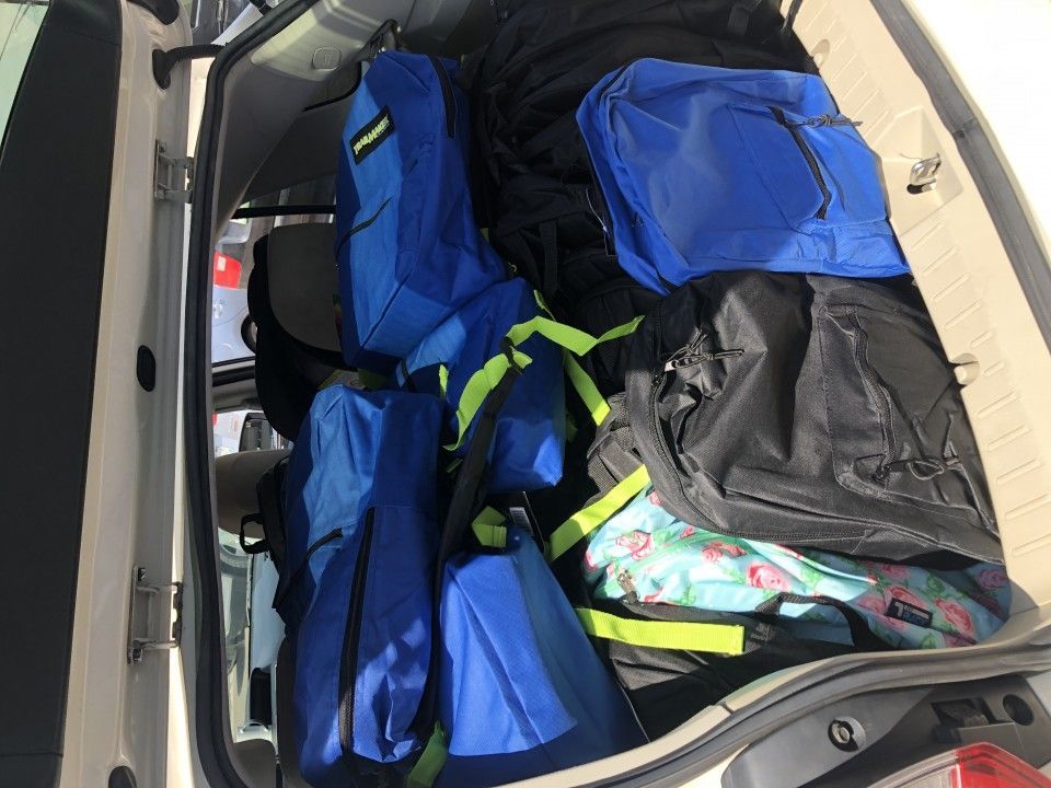 Backpack-filled car trunk; blue and black bags, some with green accents, packed inside a white vehicle.