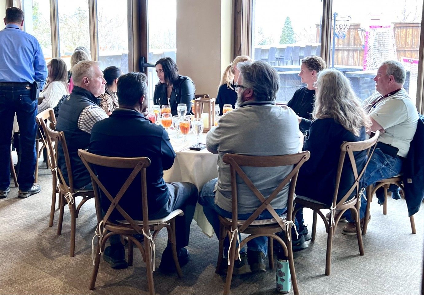 People seated at a round table, indoors. Several are facing the center; some have drinks. Soft natural light, neutral colors.