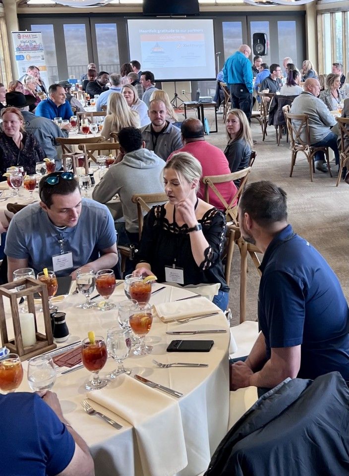 People seated at tables in a conference room, some talking, drinks and a screen in the background.