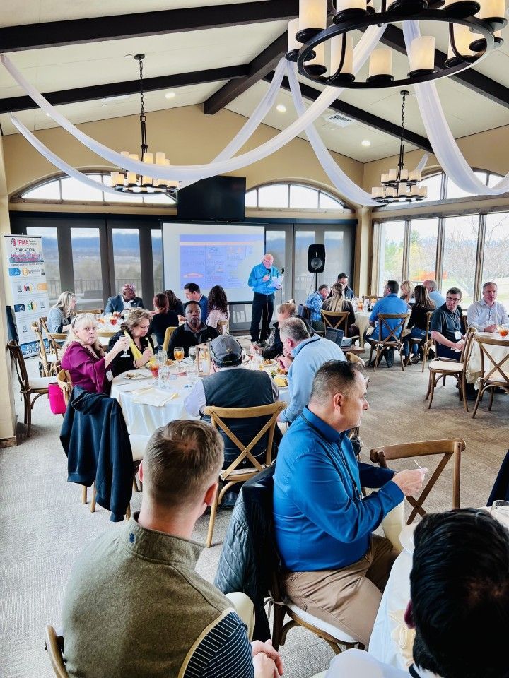 People at a conference in a dining hall, listening to a speaker. Tables are set for a meal.
