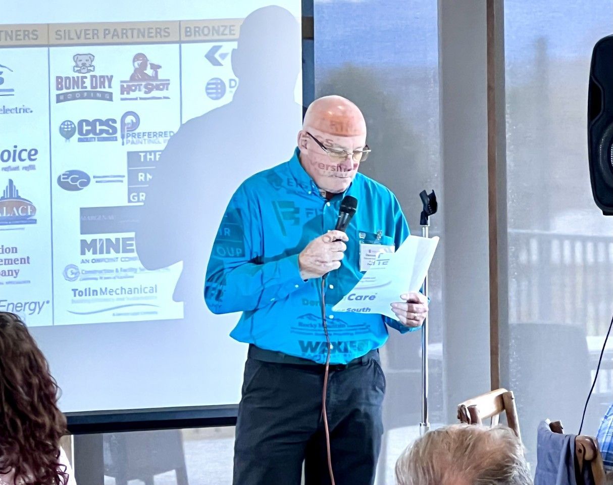 Man in blue shirt speaks into microphone, reading from papers, at a podium near a projector screen.
