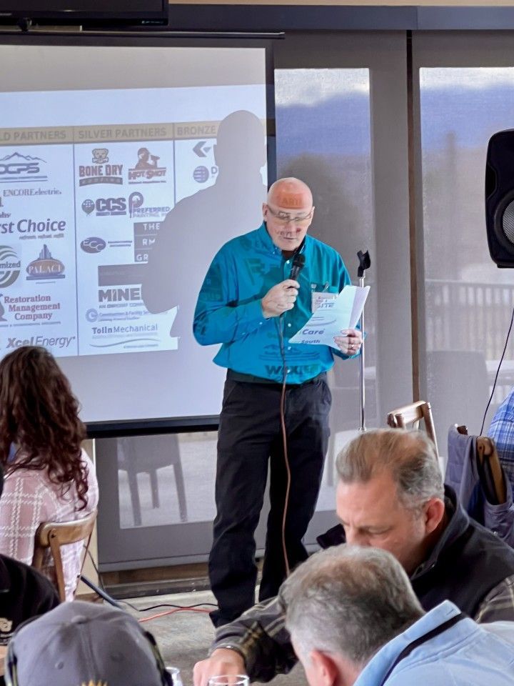 Man speaking at a conference, holding a paper and microphone, in front of a projector screen with logos.