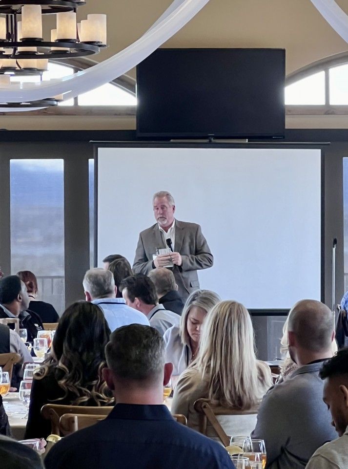 Man speaking at event, standing in front of a projection screen; audience seated at tables.