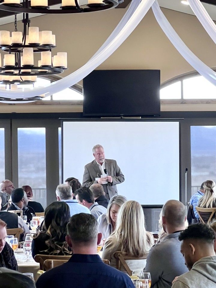 Man speaking at a conference. Audience sits at tables. Screen displays blank presentation. Indoor setting.