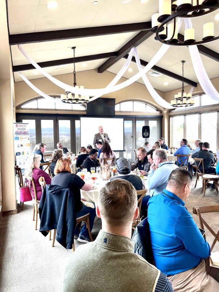 People seated at round tables listening to a speaker in a room with large windows, chandeliers, and draped ceiling fabric.