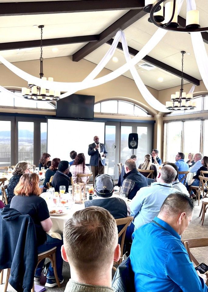 Man speaking at an event in a dining room with a crowd seated at tables. Decorative ceiling draping.