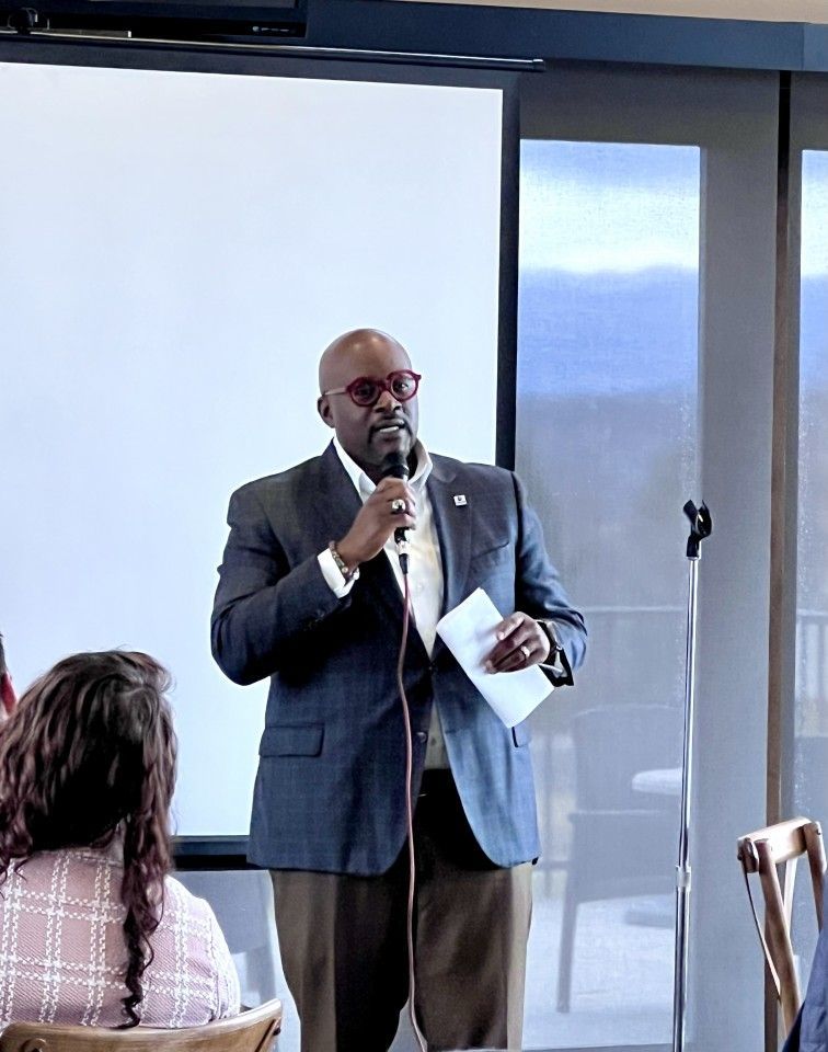Man in a blazer speaks into a microphone, holding papers, in front of a screen.