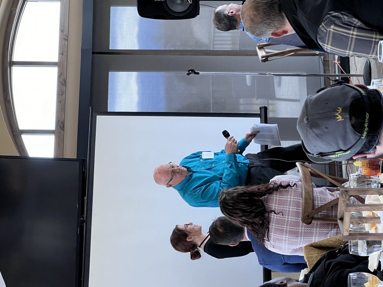 A speaker with a microphone addressing an audience in a room with a projector screen and windows.