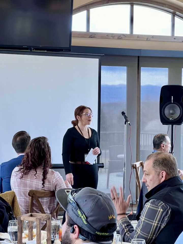 Woman presenting at a business event with a screen, microphone, and seated audience. Scenic view visible.