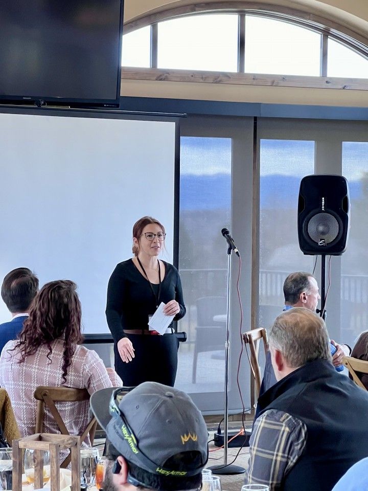 Woman speaking at a presentation, standing near a screen. Audience seated at tables in a room with a view.