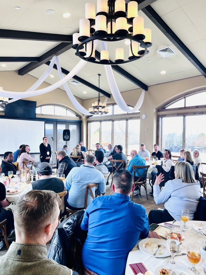 A group of people at a business meeting; a woman is speaking at the front with a projector screen.