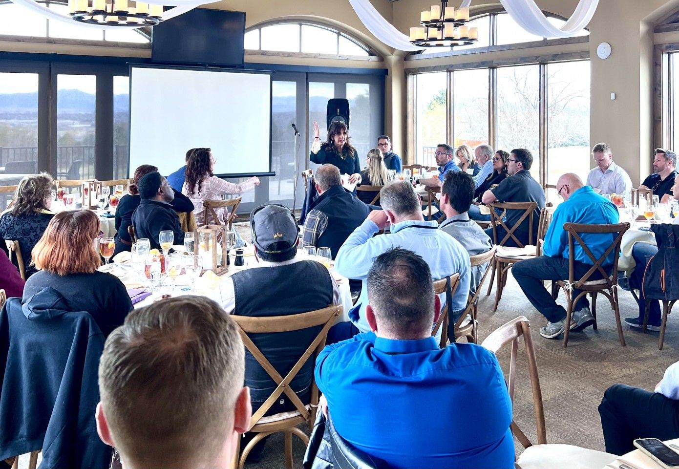 People seated at tables in a room, watching a presentation on a screen. Large windows show an outdoor view.