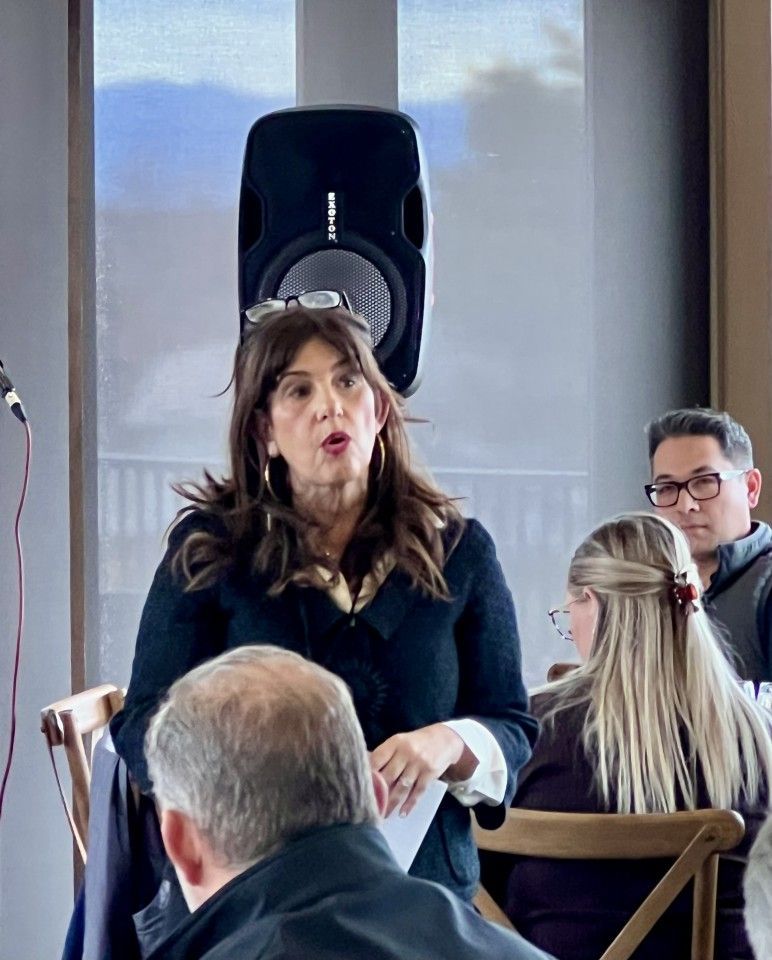Woman speaking at event; microphone, speaker, and audience visible.