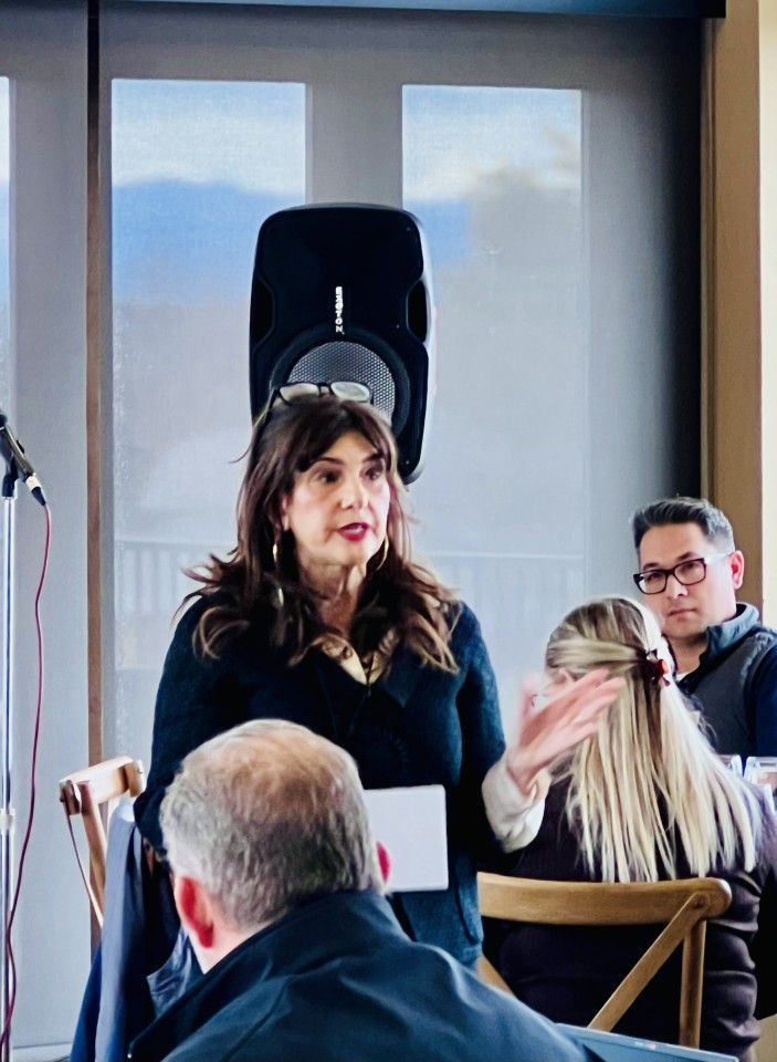 Woman speaking at a table, holding paper. Others listen at tables, indoor setting with speaker.