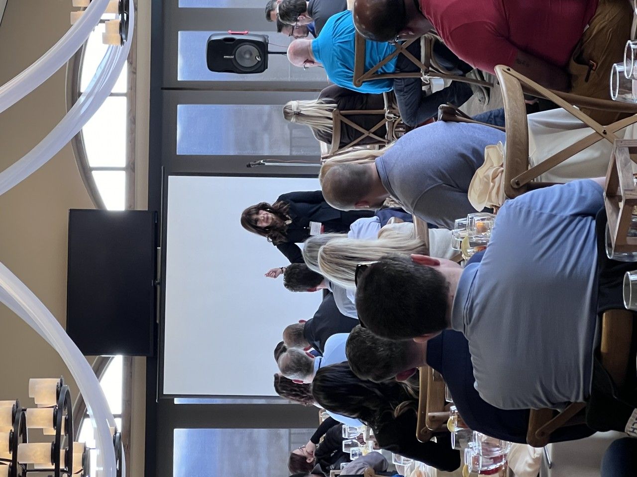 People seated at tables, listening to a presentation on a screen. A woman gestures in front.