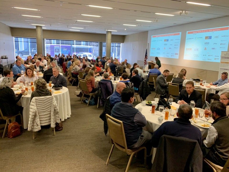 Large room with round tables, people seated, eating and talking. Two projected screens in the back.
