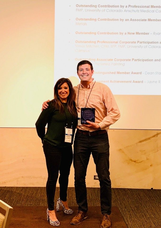 Two people smiling, one holding an award plaque; on a stage in front of a projection screen.