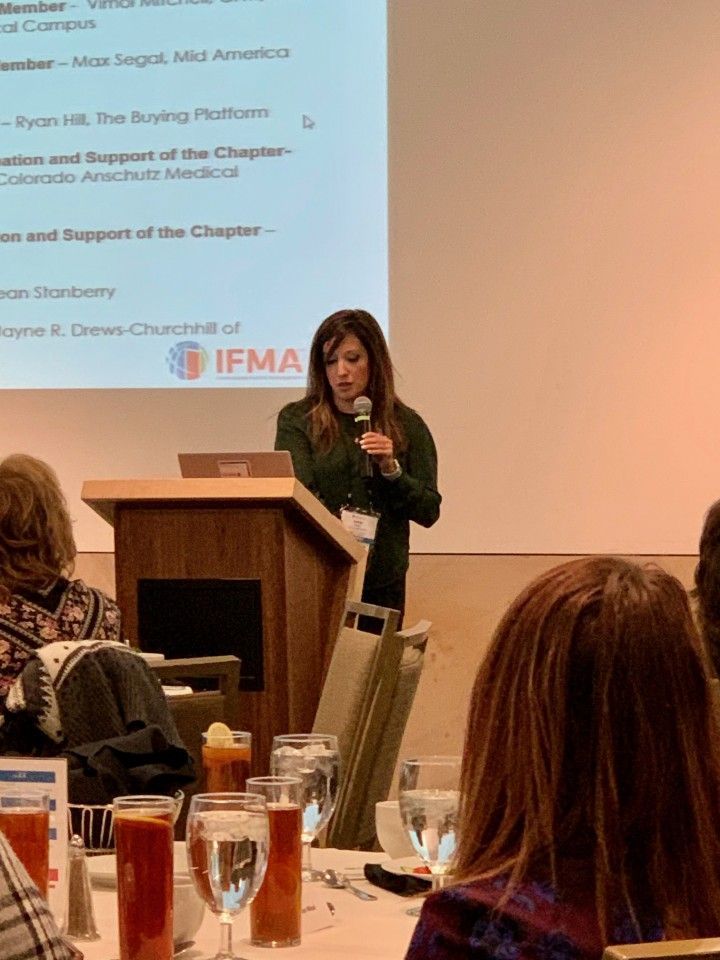 Woman speaking at a podium with a presentation screen visible. Attendees seated at tables.