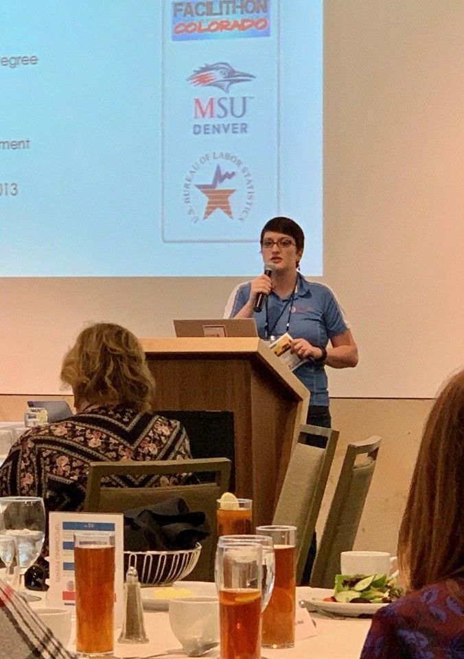Person speaking at a podium during a conference. Audience seated at tables, a slide with logos in the background.
