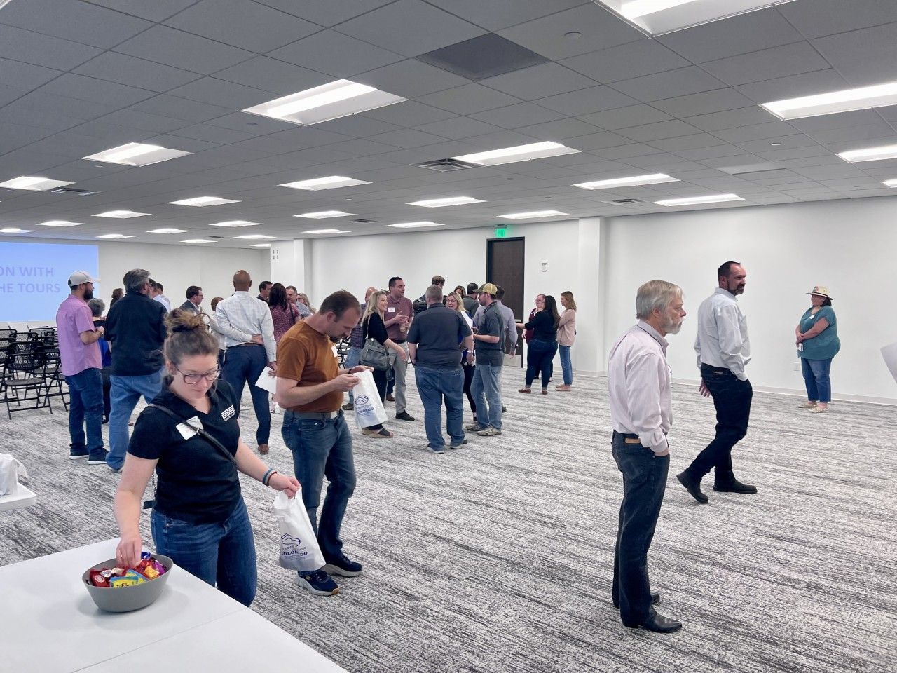 People at an event, mingling in a large room. Some are holding papers or food, others are talking. Bright overhead lights.
