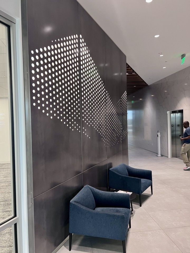 Modern office reception area with perforated dark wall art, blue armchairs, and hallway with elevator.